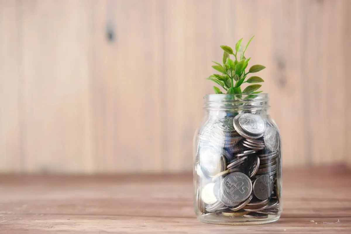 Un tarro de cristal lleno de monedas con una pequeña planta creciendo, símbolo de hacer crecer el dinero al invertir