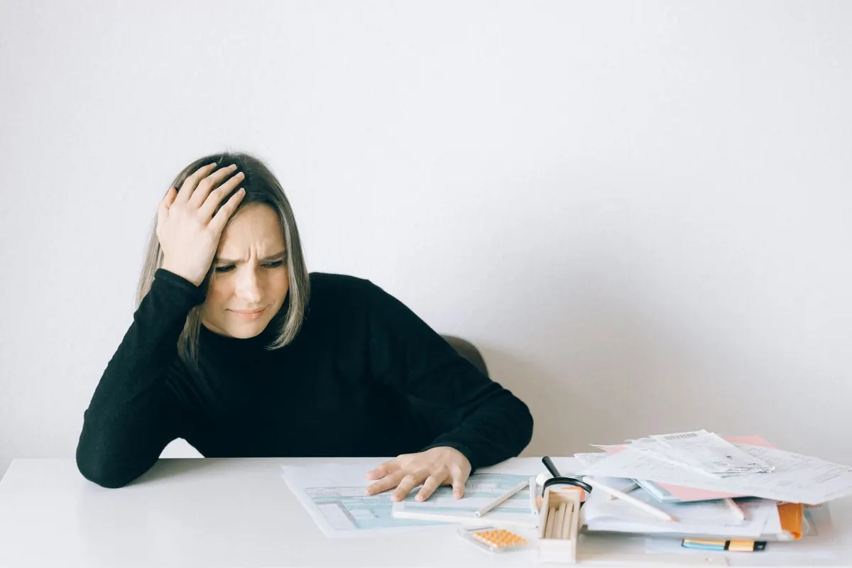 A woman resting her hand on her head while looking at a pile of bills and paperwork