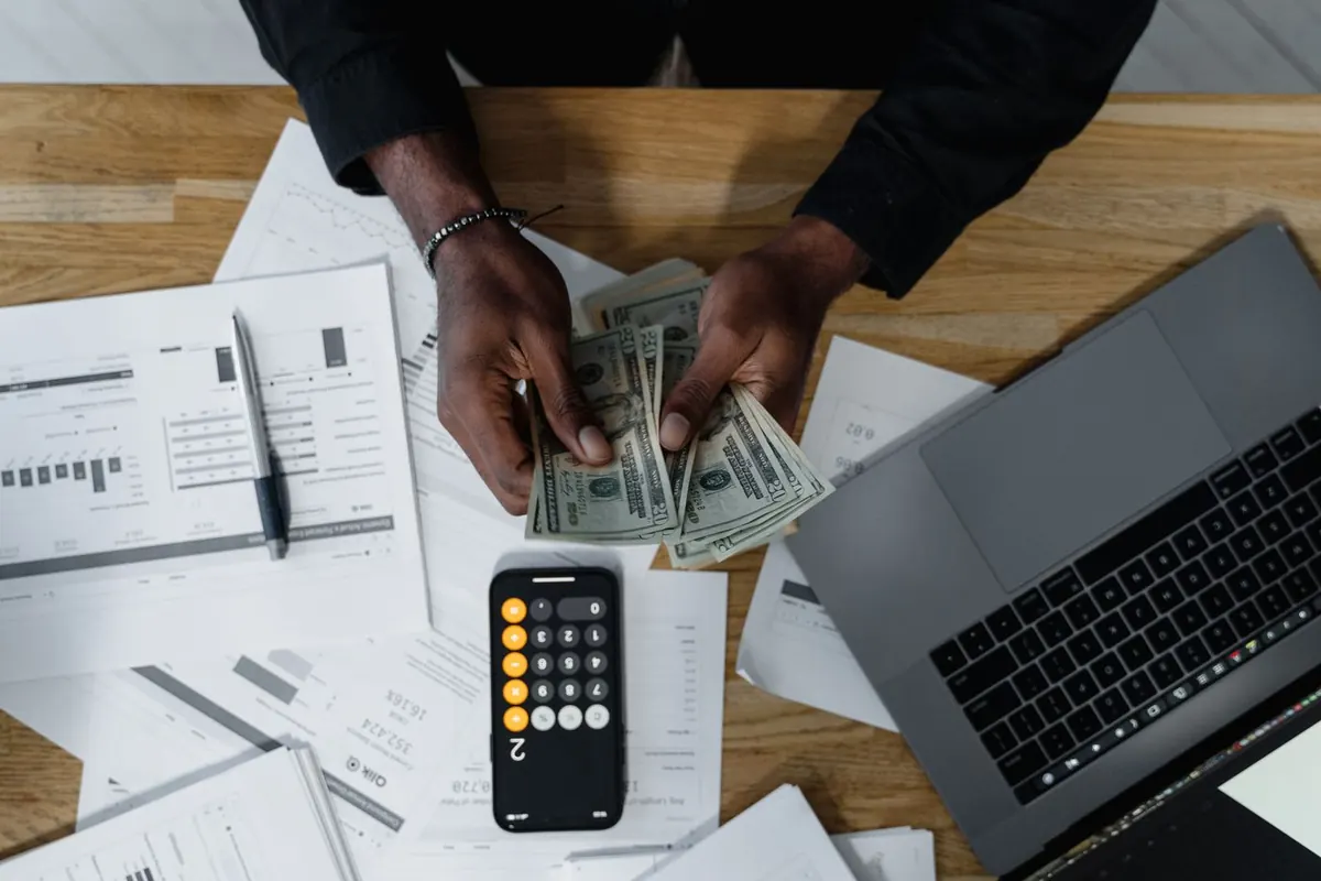 Vista cenital de unas manos contando billetes sobre documentos financieros, junto a una calculadora en el móvil y un portátil