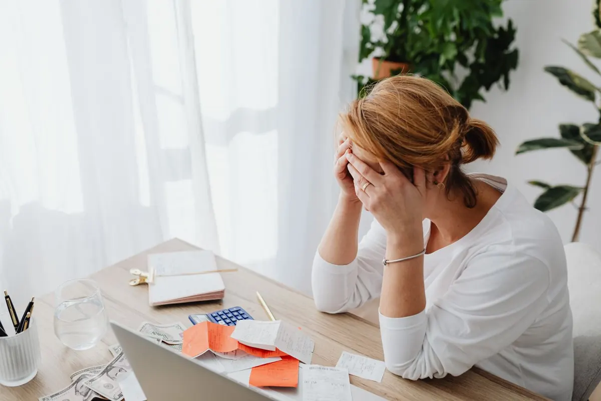 Femme assise à son bureau, la tête dans les mains, entourée de factures, d'une calculatrice et de billets