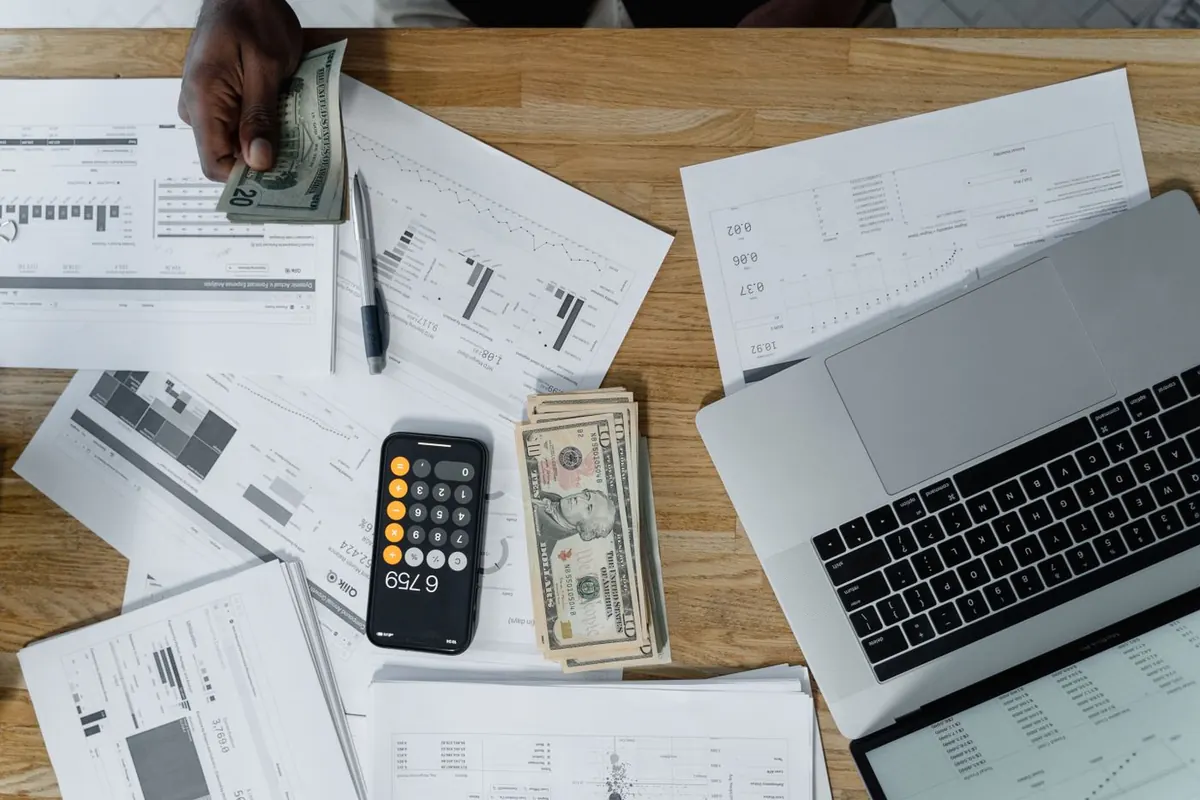 Overhead view of a desk with cash, financial statements, a phone calculator and a laptop